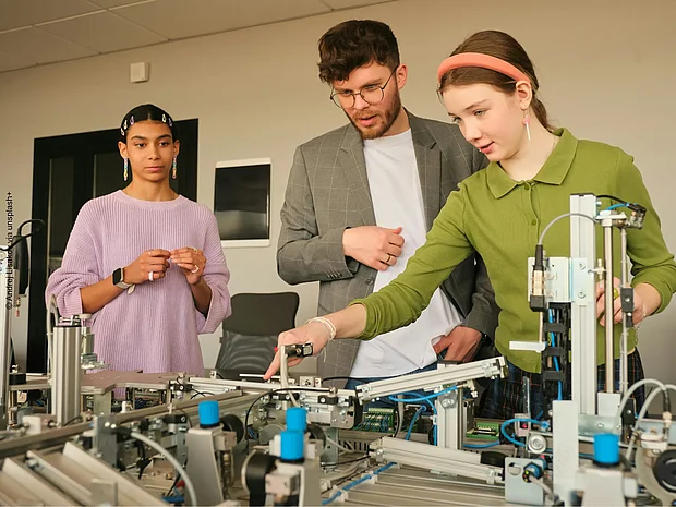 Lehrkraft zeigt zwei Schüler*innen wie sie einen physikalischen Versuchsaufbau bedienen können.