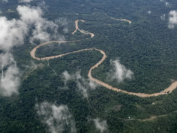 Unter einzelnen Wolken schlängelt sich ein Fluss inmitten eines grünen Regenwaldmeeres. Klitzeklein sin einzelne Straßen und Plätze zu erahnen.