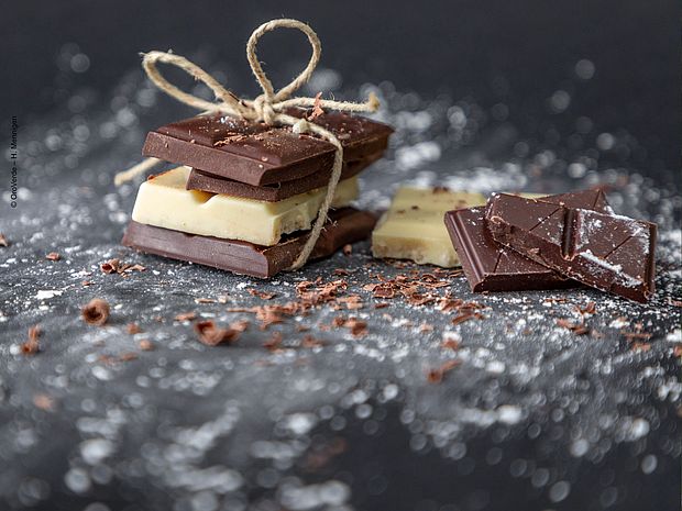 Auf einem schwarzen Hintergrund liegt ein Päckchen mit drei Schokoladenstückchen. Es hat eine goldene Schleife und ist mit Puderzucker verziert. Das Bild ist künstlerisch und hochwertig.