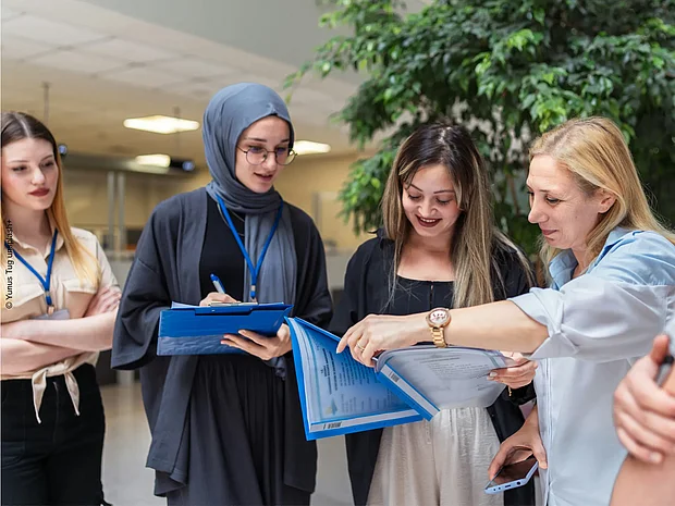 Lehrerin berät drei Schüler*innen und zeigt dabei auf die Aufzeichnungen einer Schüler*in