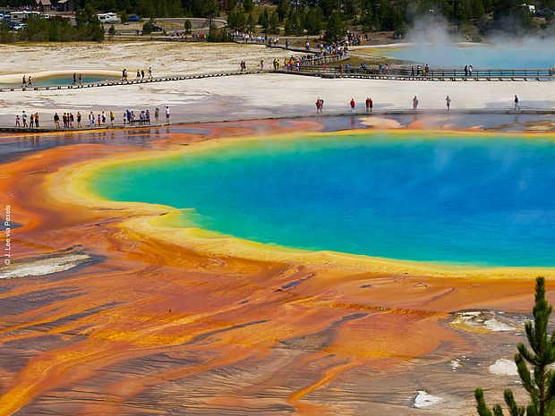 Bunte heiße Quelle im Yellowstone-Nationalpark mit türkisblauem Wasser, orangefarbenen Ablagerungen und vielen Besucherinnen und Besuchern auf Holzwegen.