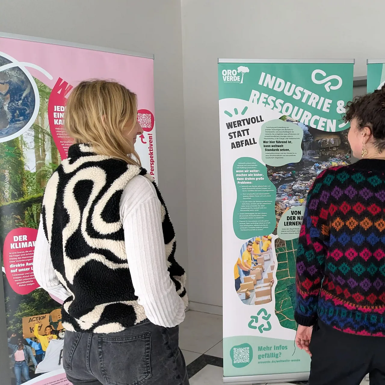 Zwei Personen stehen mit dem Rücken zu uns vor zwei große Postern. Auf einem Poster steht "Industrie und Ressourcen". Die Poster gehören zur Ausstellung "Weltweite Wende" von OroVerde