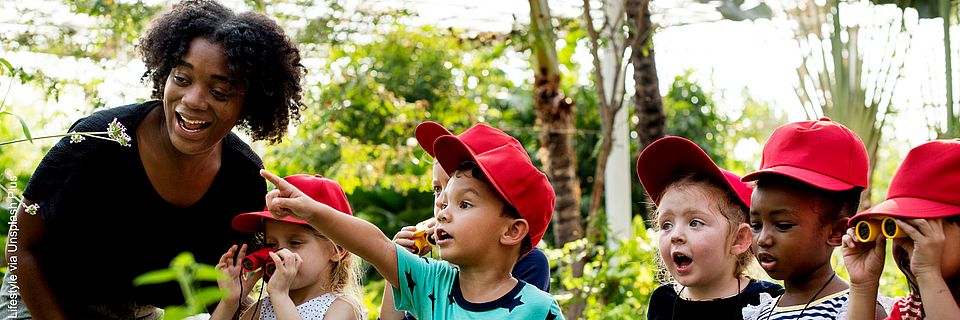 Eine Gruppe Kinder und ihre Lehrkraft stehen in einem großen Gewächshaus. Ein Kind zeigt aufgeregt in die Ferne. Zwei Kinder schauen durch Ferngläser in dieselbe Richtung.