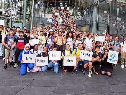 ine große Gruppe von Menschen, darunter Kinder und Erwachsene, steht auf den Stufen eines Glasgebäudes. Einige halten Schilder mit der Aufschrift „Ein Engel für den Wald“.