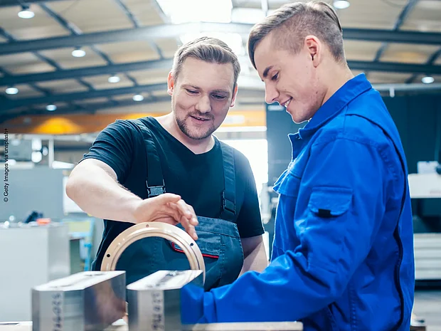 Aubilder im schwarzen T-Shirt hat seine Hand an einem Lehrstück der Metallverarbeitung und erklärt etwas. Ein Lehrling im Blaumann steht rechts von ihm und hört interessiert zu.