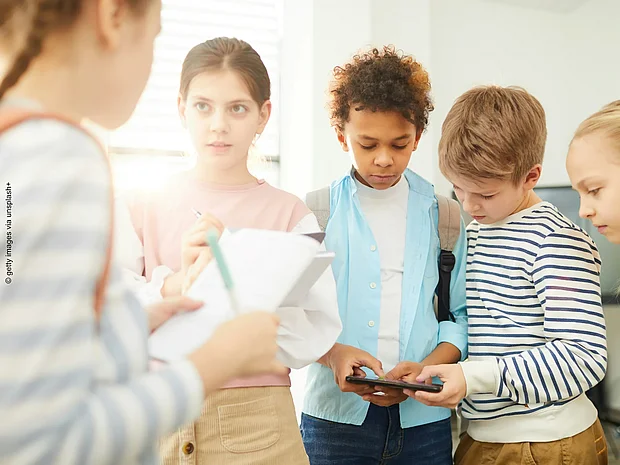 Vier Schüler*innen stehen in einem Halbkreis und schauen auf ein Tablet und auf ausgedruckte Zettel
