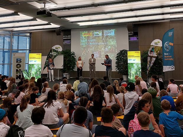 Eine Präsentation über Recyclingpapier mit einer Gruppe von Kindern im Publikum und vier Personen auf der Bühne. Im Hintergrund ist eine Leinwand mit dem Titel „Paper Angels: Wir für Recyclingpapier!“ zu sehen.