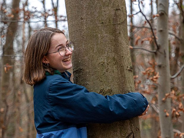 Eine junge Frau umarmt einen Baum. Sie freut sich dabei sehr.
